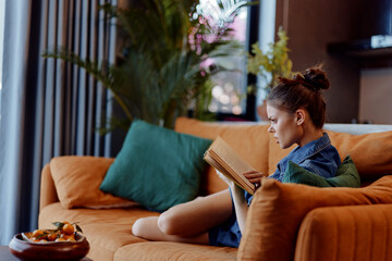Obraz premium Young woman reading a book while sitting comfortably on an orange couch at home