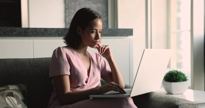 Thoughtful freelance entrepreneur woman looking away, thinking, searching for inspiration, making aha finger gesture, turning to laptop, typing, working on online business project from home couch