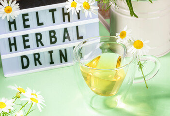 Herbal medicine. Healthy chamomile tea in glass cup, flowers, inscription by black letters on green.