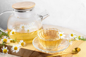 Tea drinking for health. Glass teapot and cup with chamomile tea, spoon, daisies on wooden board.