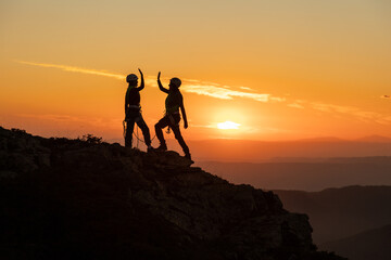 Two people are on a mountain, one of them is giving the other a high five. The sun is setting in the background, creating a beautiful and serene atmosphere