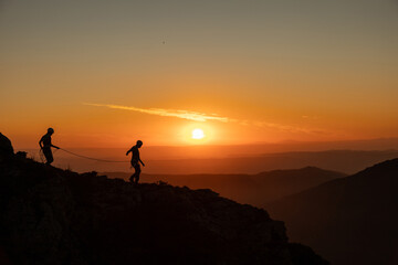 Two people are walking on a mountain side, with the sun setting in the background. Scene is peaceful and serene, as the two people enjoy the beautiful view of the sunset