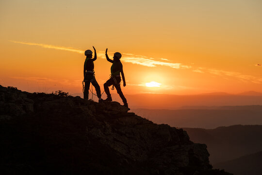 Two people are on a mountain, one of them is giving the other a high five. The sun is setting in the background, creating a beautiful and serene atmosphere