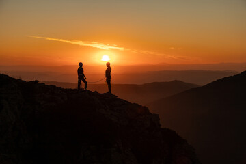 Two people are standing on a mountain top, looking out at the sunset. The sky is orange and the sun is setting in the distance