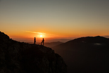 Fototapeta premium Two people are standing on a mountain top, looking out at the sunset. The sky is a mix of orange and pink hues, creating a serene and peaceful atmosphere