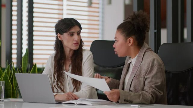 Multiracial businesswomen African American Caucasian girls displeased coworkers office company discuss documents teamwork analyze papers female business partners women disagree fight conflict quarrel