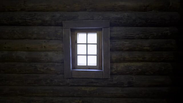 A small window in a traditional Russian wooden izba illuminates the dark log interior with soft natural light. The contrast between the warm, rough logs and the bright window creates a cozy, rustic