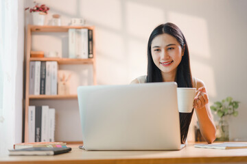 Young Asian woman using a laptop while enjoying a drink in her home office.