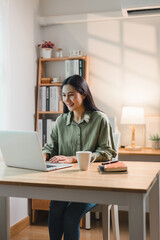 Young asian woman happily working on laptop in modern home office. The space is well-lit and features stylish decor, creating a productive atmosphere.