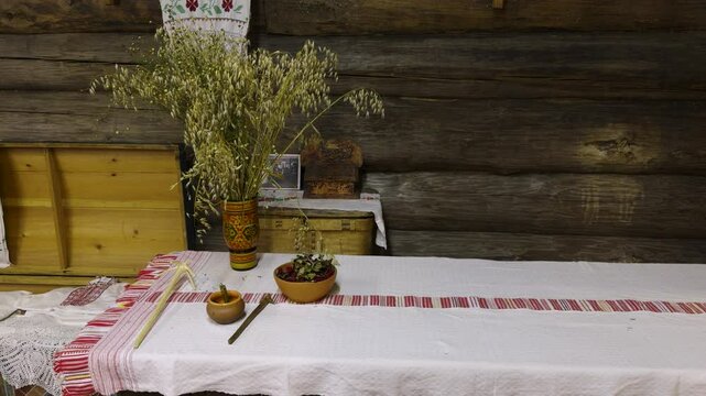 A cozy scene of a traditional table in a Russian izba. The table is covered with a white embroidered tablecloth and decorated with dried oats in a painted vase, clay bowls, and handicrafts. The rustic