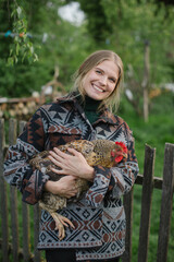 Pretty blonde farmer holding chicken in hands while standing in yard in rural area.