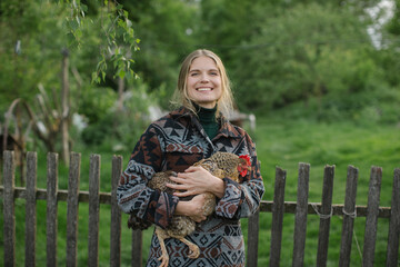 A young woman holds a chicken in her hands while standing in a farm yard in the countryside.