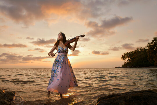 Young woman in a long dress standing in the sea playing the violin at sunset, Thailand
