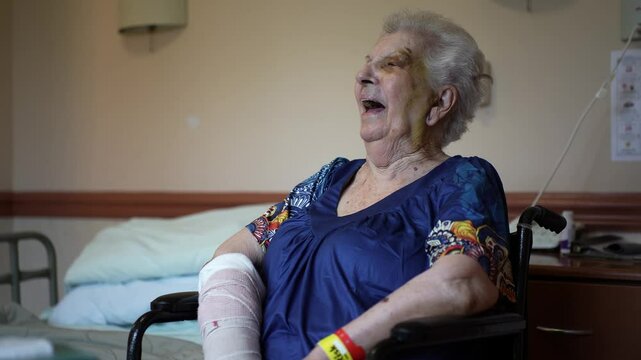 Smiling elderly woman in muumuu and cast on arm with bruised face black eye sitting in wheelchair in rehabilitation facility.