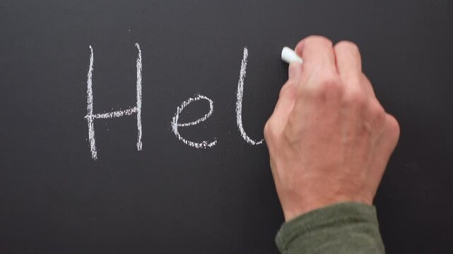 A hand writes the word hello with white chalk on a black board