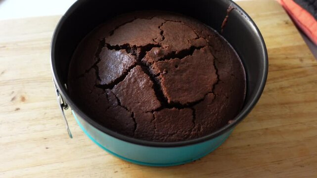 A freshly baked chocolate cake cracked in a round blue baking pan, placed on a wooden cutting board next to an induction stove and a black oven mitt, ready to be served