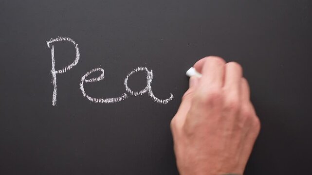 A hand writes the word peace with white chalk on a black board