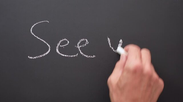 A hand writes the words or expression &ldquo;see you&rdquo; with white chalk on a black board