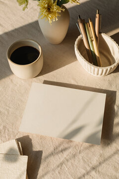 Overhead view of a blank piece of card on a table with assorted stationery, a cup of black coffee and a vase with flowers in sunlight