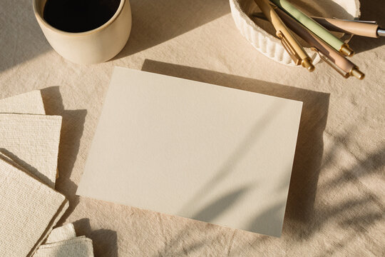 Overhead view of a blank piece of card on a table with assorted stationery, a cup of black coffee and a vase with flowers in sunlight