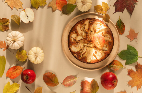 Overhead view of a homemade apple pie with fresh apples, autumn leaves, viburnum twigs and decorative white pumpkins on a beige background
