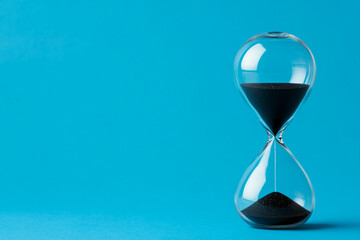 A glass hourglass with sand in it is on a blue background. Concept of time passing quickly and the fleeting nature of time