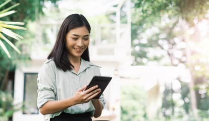 Asian business woman working outside using portable tablet device wireless remote communication technology, connectivity digital media online freelance worker outdoor in nature tree at cafe sunlight.