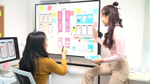 Two asian ui/ux designers are discussing user interface of mobile application prototype on whiteboard, collaborating on user experience design