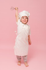 A young baby boy wearing a baker's hat and apron is captured in a studio shot, holding a whisk, with a pink background setting. Kid aged 3 years (three year old boy)