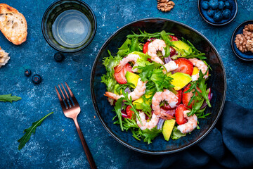 Fototapeta premium Foodie food shrimp salad with strawberry, arugula, lettuce, avocado and red onion, blue background