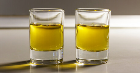 Two shot glasses filled with golden liquid, likely oil or alcohol, placed side by side on reflective surface.
