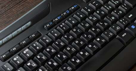 Black keyboard with dual language Cyrillic and English layout on wooden desk, featuring function...