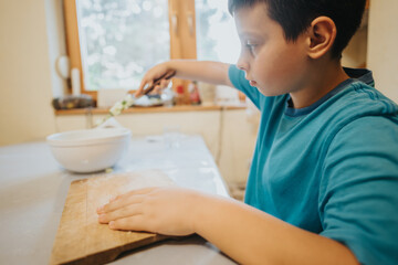 A young boy in a blue shirt concentrates on preparing food in a bright kitchen, showcasing focus and learning through a cooking activity.