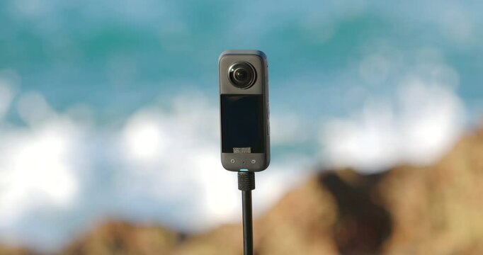 Close-up of 360 camera stands on tripod near ocean waves. .