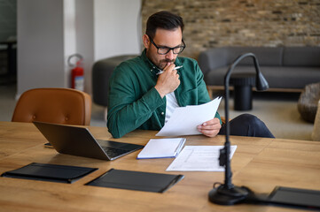 Focused male entrepreneur in eyeglasses analyzing business report while working at desk in office