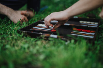 Close-up of hands playing backgammon on a grassy field, reflecting leisure and strategy. The image captures a tactile moment in a social outdoor setting.