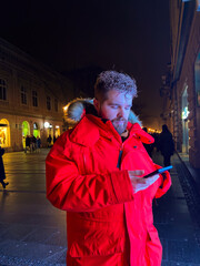 Millennial man in vivid red parka stands on city street at night, engrossed in their smartphone....
