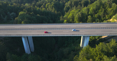 AERIAL: Two cars speed along the modern freeway bridge above the dark green forest. Flying over the...