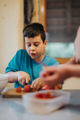 A young boy is focused on learning to cook, slicing vegetables on a kitchen table. The scene captures concentration, skill development, and a love for cooking.