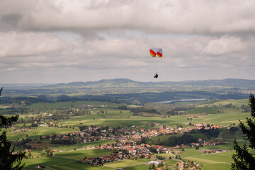A paraglider soars over a scenic Bavarian village with rolling hills, green fields, and scattered...