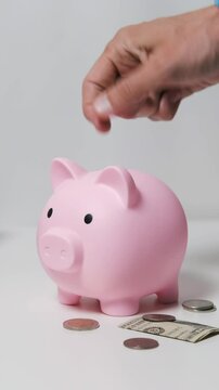 Vertical video. Coin drops into piggy bank among scattered coins and a dollar, with books and a notebook in the backdrop