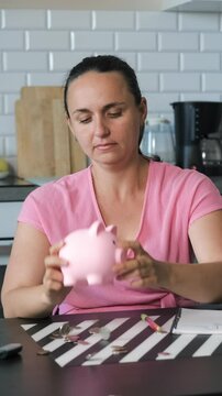 Vertical video. Young woman pours money from piggy bank, coins scatter across a kitchen table