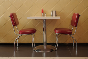 Fototapeta premium Empty booth at retro-style diner with red upholstered chairs and white table adorned with condiment set against yellow quilted wall