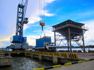 A heavy-duty industrial crane lifting a large blue shipping container at a busy port, set against a backdrop of a bright blue sky and ocean. The scene highlights the efficient cargo handling process