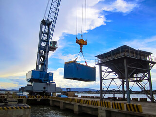 A heavy-duty industrial crane lifting a large blue shipping container at a busy port, set against a backdrop of a bright blue sky and ocean. The scene highlights the efficient cargo handling process