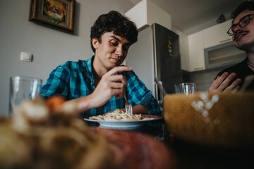 Two friends share a relaxed meal at home, enjoying pasta and conversation in a cozy dining area with a modern kitchen backdrop.