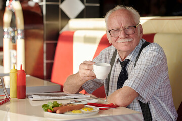 Portrait of elderly man with mustache sitting in diner, smiling while holding coffee cup, with food on table and red, white booth in background