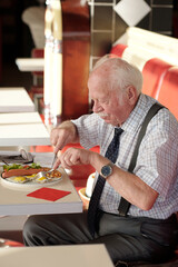 Elderly man enjoying meal while seated in cozy restaurant setting. Dressed in formal attire and using utensils, he is concentrating on his food without distractions