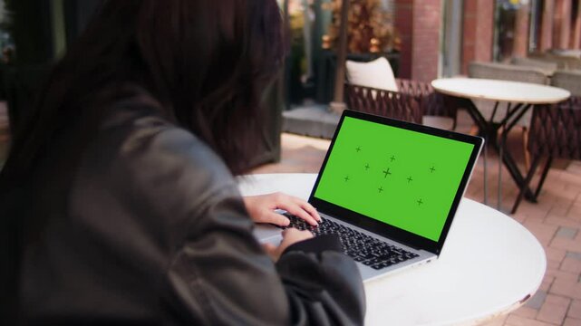 Young girl watching on a laptop with a mockup green screen. Woman in a black jacket is sitting at the outdoor cafe and scrolling feed on a computer, back view