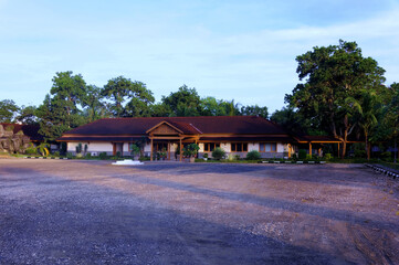 A serene villa surrounded by lush greenery, featuring a traditional yet modern architectural design with a sloping red-tiled roof. The villa is nestled amidst a garden landscape